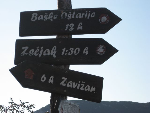 Kažipot na ovinku ceste desno nad kočo, pri partizanskem spomeniku